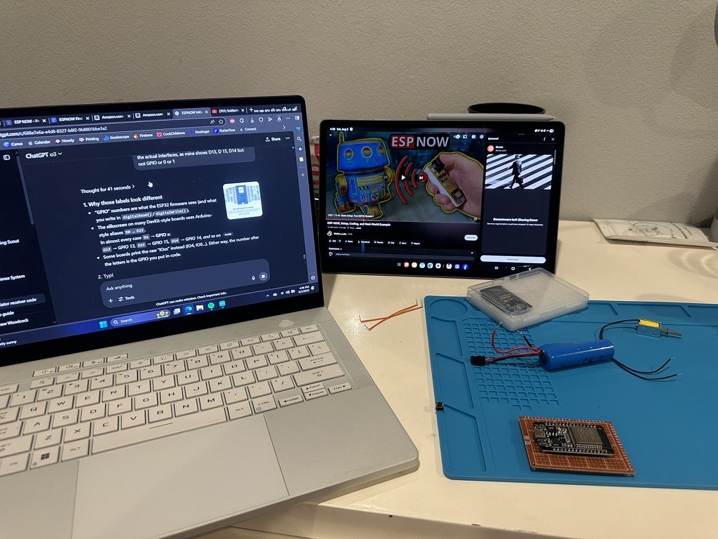 Electronics prototyping bench with an ESP32 board, battery, soldering mat, laptop, and ESP-NOW reference material.